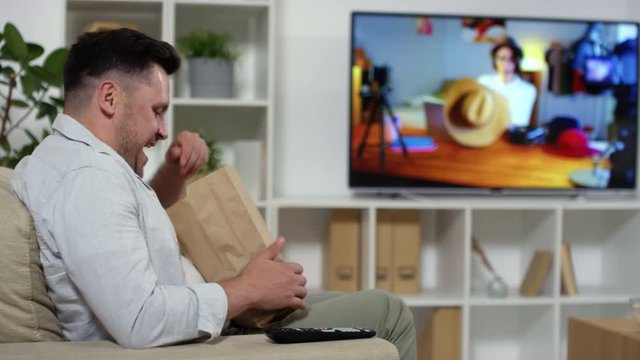 Cheerful Caucasian man bringing coffee cups on tray and street food in living room. He sitting in armchair, opening box and enjoying smell of nuggets before eating dinner in front of TV