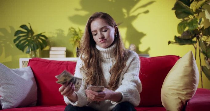 Young red-haired business woman receiving salary counting cash money rejecting low profits showing unsatisfaction. Home interior.