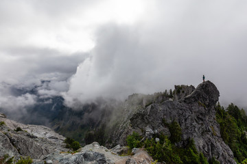 Fototapeta na wymiar Adventurous Man Standing on top of a rugged rocky mountain during a cloudy summer morning. Taken on Crown Mountain, North Vancouver, BC, Canada.