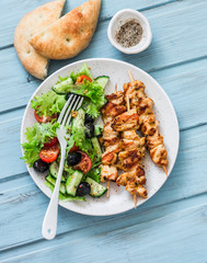 Chicken souvlaki kebabs, sun dried olives, tomatoes, cucumbers green salad and focaccia on blue...