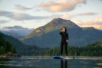 Adventurous Girl on a Paddle Board is paddling in a calm lake with mountains in the background during a colorful summer sunset. Taken in Stave Lake near Vancouver, BC, Canada.