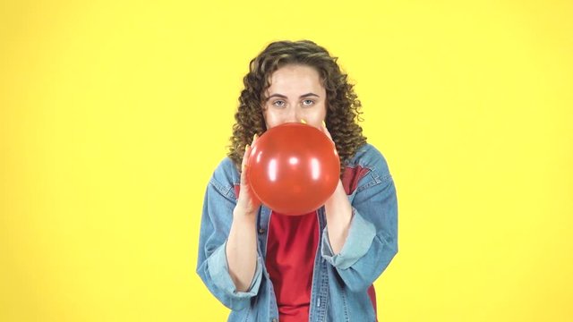 Curly girl inflates a red ball then blows it off on yellow background, slow motion.