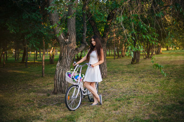 Fototapeta na wymiar girl in a white skirt and a vest with a bicycle and a basket full of flowers in the park in the evening