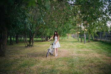 Fototapeta premium girl in a white skirt and a vest with a bicycle and a basket full of flowers in the park in the evening