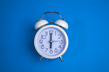 A white alarm clock on a blue background. a top view. a close-up picture representing 6 o'clock