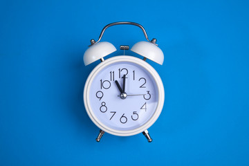 A white alarm clock on a blue background. a top view. a close-up picture representing 11 o'clock