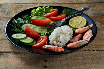 Rustic style. Shrimp with rice and vegetables (tomatoes, cucumbers, lemon, greens) on a black plate.