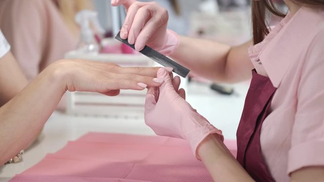 A manicurist wears the pink gloves. A table is white. The girl uses nail strip.