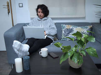 Young caucasian woman working on her laptop with her feet on the table, in pajamas, sitting on the sofa with a cup of coffee