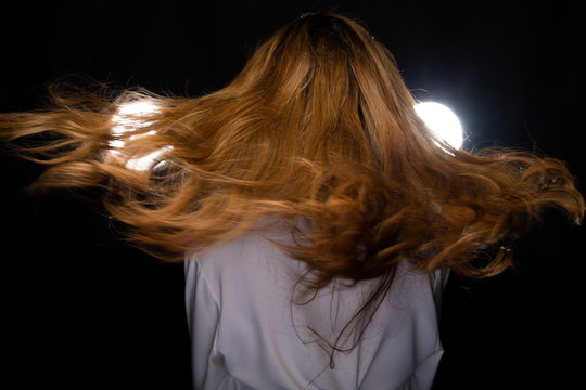 Girl is flying and throwing windy curl hair around.  Beautiful Asian Woman flutters blonde Brown dying color hair style in the air and spin around over dark background with two backlit, back side view