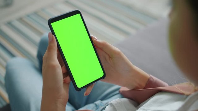 Female hand is holding big modern smartphone with blank green screen without touching it sitting at home using device. Technology, communication and equipment concept.
