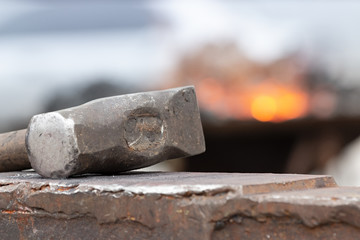 Old rusty hammer lies on the anvil with flame of brazier forge on background. Blacksmith, metalsmith, farrier tools. Close view.
