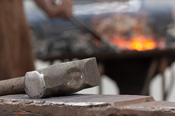 Old rusty hammer lies on the anvil with blacksmith work at the brazier forge on background. Blacksmith, metalsmith, farrier tools.