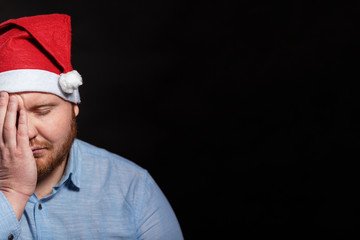 A sad man with a red beard in a Santa Claus hat covered his face with his hand. Black background....