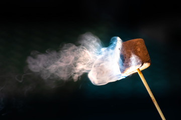 Roasting chocolate coated marshmallow on a stick; smoke swirls visible on a dark background