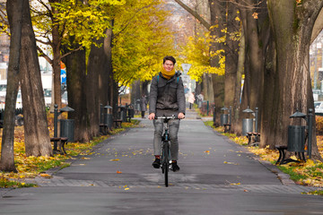 tall smiling guy of European appearance in glasses, with a yellow scarf and a gray jacket rides a Bicycle along the Avenue of the autumn Park. wet path after rain, fallen yellow leaves