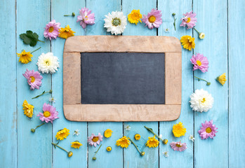 Blackborad frame on center of fresh flowers with leaves on light blue wooden background. Flowers composition. Flat lay. Top view. Copy space on blackboard.