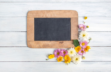 Blackborad frame with fresh flowers and leaves on lower right corner on white wooden background. Flowers composition. Flat lay. Top view. Copy space on blackboard.