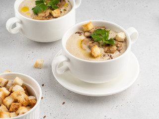 Delicious and creamy mushroom (champignon) cream soup in white ceramic bowls with green fresh parsley, flax seeds and  crunchy croutons. White concrete background
