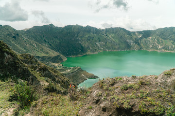 Fototapeta na wymiar Volcano crater lake view, Quilotoa. Dramatic perspective of Quilotoa lake and volcano crater, with view of mountains, hiking path trail loop and cloudy sky from viewpoint. Shot in Ecuador. Green blue
