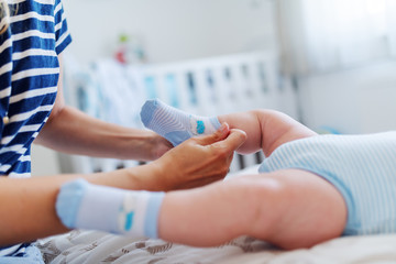 Close up of caring caucasian mother putting tiny socks on baby's feet. Baby lying on bed. Selective focus on foot.