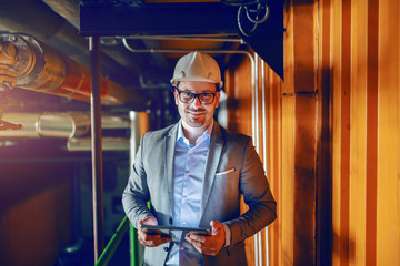 Manager in suit and with helmet on head standing in factory and holding tablet while looking at camera.