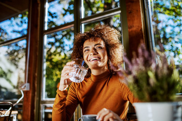 Cute smiling young mixed race woman with curly hair and in turtleneck sitting in cafe and drinking water. Morning time.