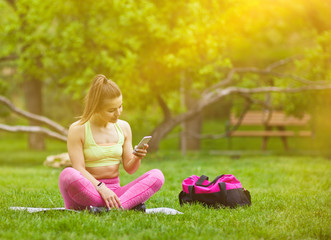 Young fit woman chatting on smartphone with friends while sitting on mat in park outdoors