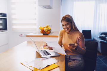 Cute caucasian smiling blonde woman in sweater holding bills in one hand and in other smart phone. On table are laptop and bills. Apartment interior. - obrazy, fototapety, plakaty
