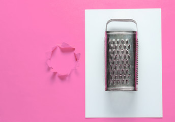 Old metal grater on pink paper background with torn hole for copy space. Top view. Studio shot