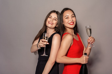 Freestyle. Young girls in dresses standing isolated on gray with champagne and glasses posing smiling happy