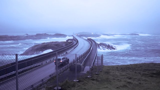 Slow motion shot of car driving over a long bridge that goes over a restless sea during stormy weather.
