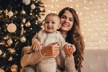 Happy family of mom with little baby hugging and celebrating new year in front of Christmas tree in decorated interior. Celebrating Christmas.