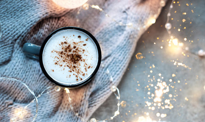 Cup of coffee with cinnamon. Christmas background with lights, warm knitted scarf.