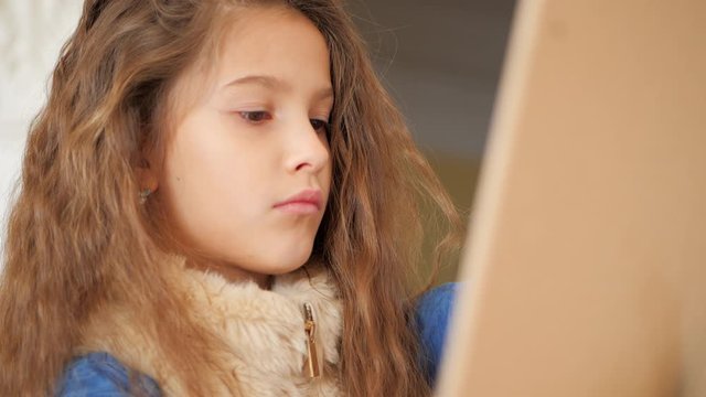 Portrait. A young girl with curly blond hair draws with a pencil at the easel. Dolly zoom