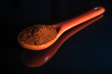 Small orange ceramic spoon with chili powder spice, with reflection on shiny black background