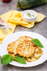 Vegetable fried fritters from zucchini and basil leaves on a plate and sour cream sauce with greens in a bowl on the table