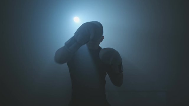 Kickboxer training in low light gym. Sportsman boxing in camera. Fighter punching. Silhouette on dark background. Close-up of boxer practicing boxing punches looking at camera in smoky gym. 4K, UHD