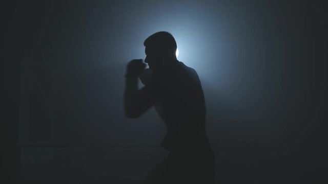 Male boxer training with punching bag on black smoky background with light. Shadow fight. Strength and motivation. Silhouette of boxer preparing for big fight. Medium shot