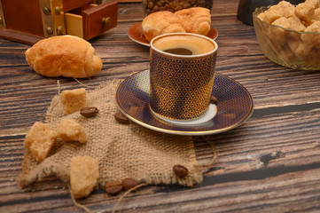 A Cup of coffee, pieces of brown sugar in a sugar bowl and croissants on a wooden background.