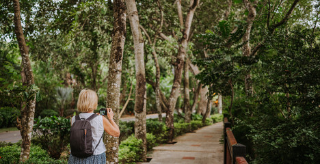 Naklejka na ściany i meble Traveler elderly senior backpacker woman walking taking photos in tropical park, Travel adventure nature in China, Tourist beautiful destination Asia, Summer holiday vacation trip, Copy space