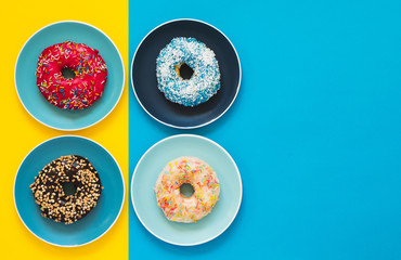 Four appetizing donuts lie on color ceramic plates on blue and yellow background with copy space.