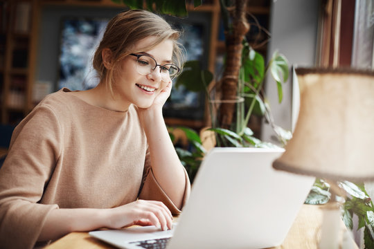 Cheerful young caucasian girl in glasses using laptop, shopping online or browsing internet, chatting with friends, edit photos for university project, smiling as looking computer screen