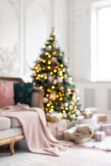 Blured photo of decorated wit garland and toys christmas tree standing next to white sofa.