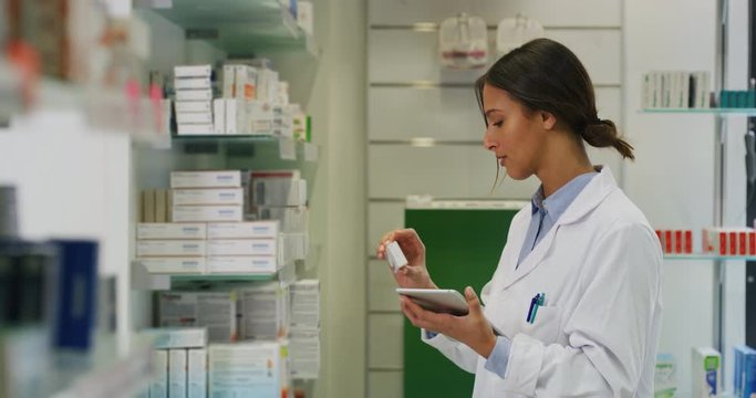 An young female pharmacist consultant is checking with a tablet medicines on the shelf of drug store and smiling in camera.