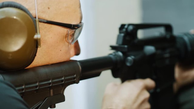 A man shoots with an automatic carbine at a shooting range, he is wearing safety eyeglasses and headphones, takes aim at a target, firing training in the shooting gallery. Close-up 4k slow-motion