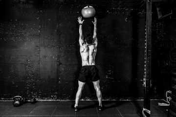 Young strong sweaty focused fit muscular man with big muscles doing throwing medicine ball up on the wall for cross training hard core workout in the gym real people selective focus black and white