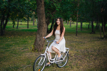 Fototapeta na wymiar A girl in a white dress rides a bicycle through the park in the evening.