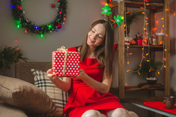 Woman in red dress sits on a sofa with a gift box in her hands on background of garland lights. Cozy Christmas mood