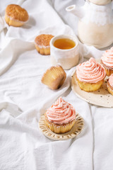 Homemade cupcakes with pink buttercream and coconut flakes served with ceramic teapot, cup of tea on white folded tablecloth.